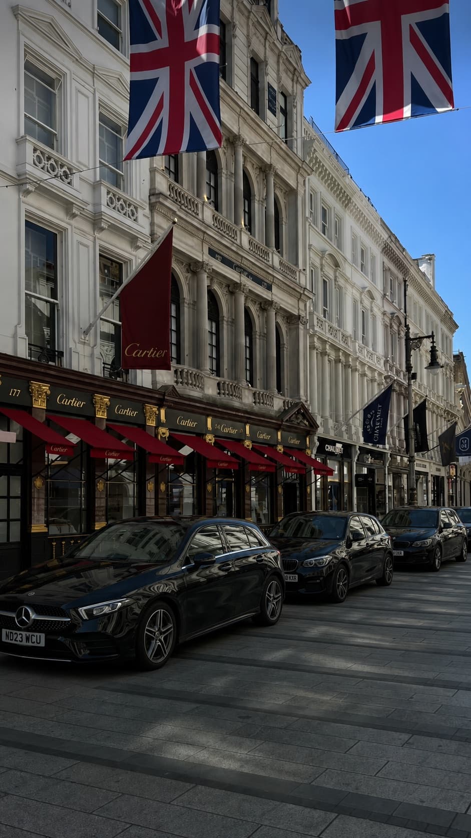 A black Mercedes-Benz A-Class and BMW 1 Series parked on a London street lined with luxury shopfronts